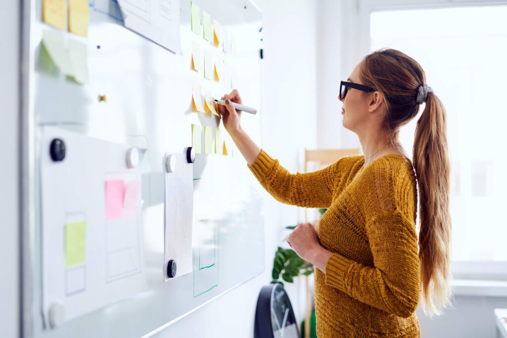 Woman writing on white board that is covered in sticky notes. Identifying knowledge gaps.