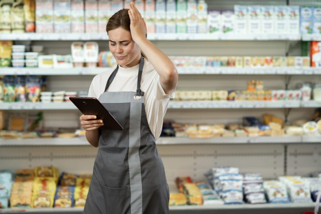 Stressed Woman Wearing Uniform With Apron Working In Supermarket Holding Digital Tablet Looking At Screen, Take Head, Grocery Shelves On Background.