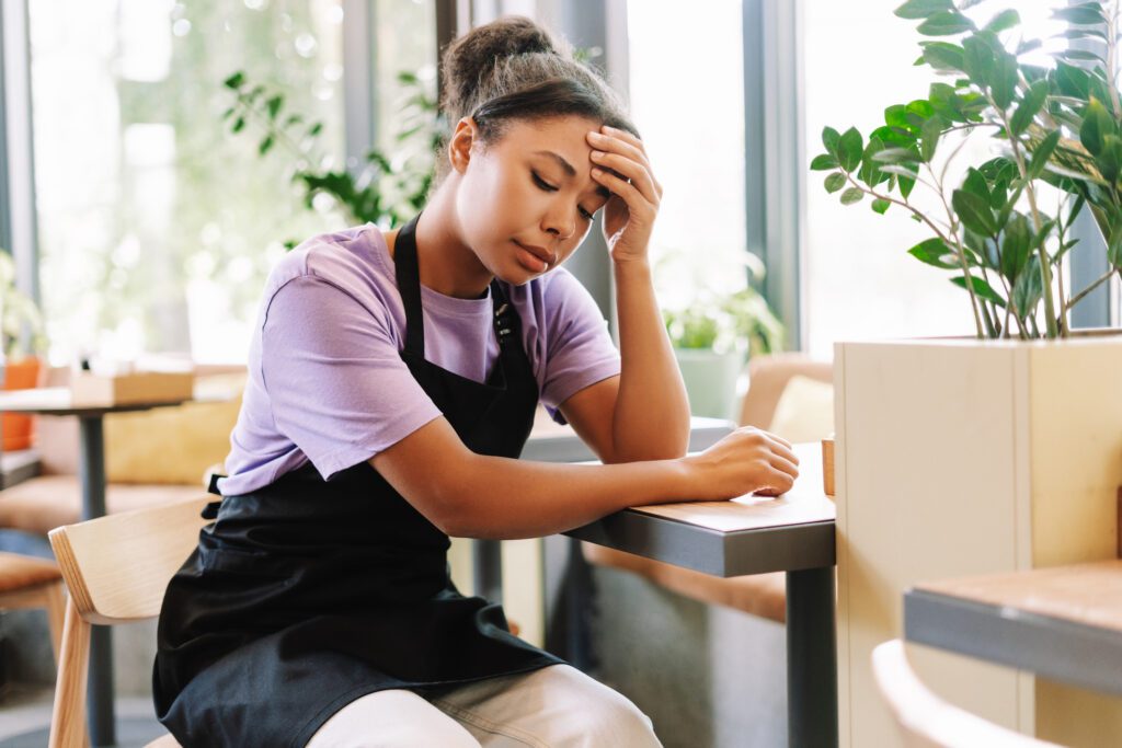 A woman employee at a restaurant sitting down at a table with her hand on her head, looking defeated.