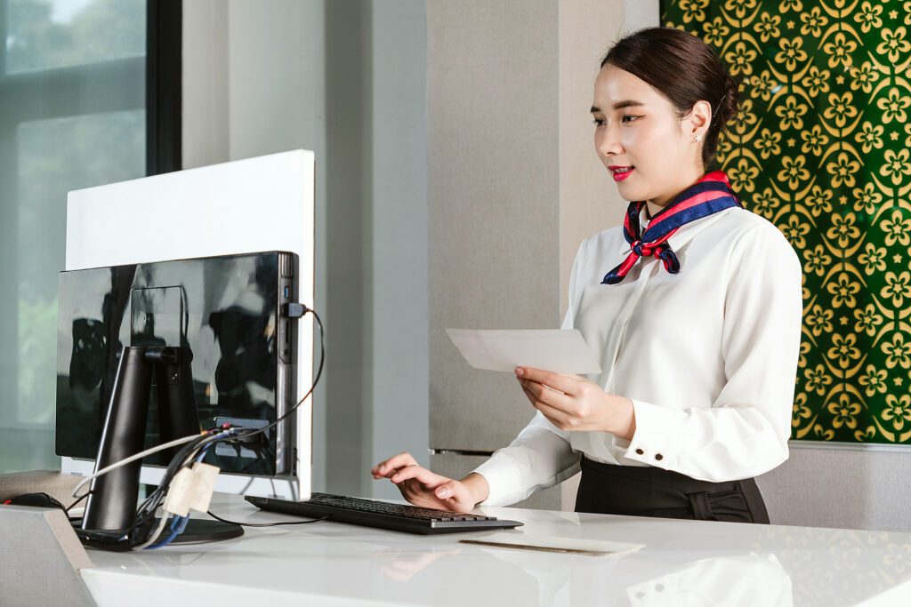 Airline Ground Staff Woman In Uniform Assists Check In At Airport Counter, Holding Ticket And Using Computer. Concept Of Air Travel Service, Passenger Support, And Airport Operations.