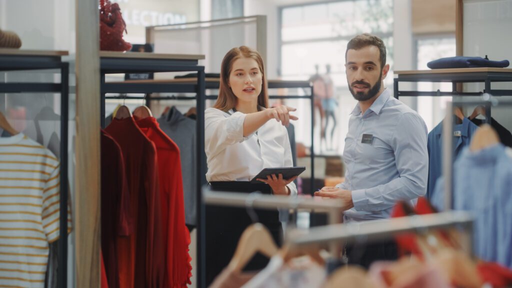 Clothing Store: Businesswoman Uses Tablet Computer, Talks To Visual Merchandising Specialist, Collaborate To Create Stylish Collection. Small Business Fashion Shop Sales Manager Talks To Designer