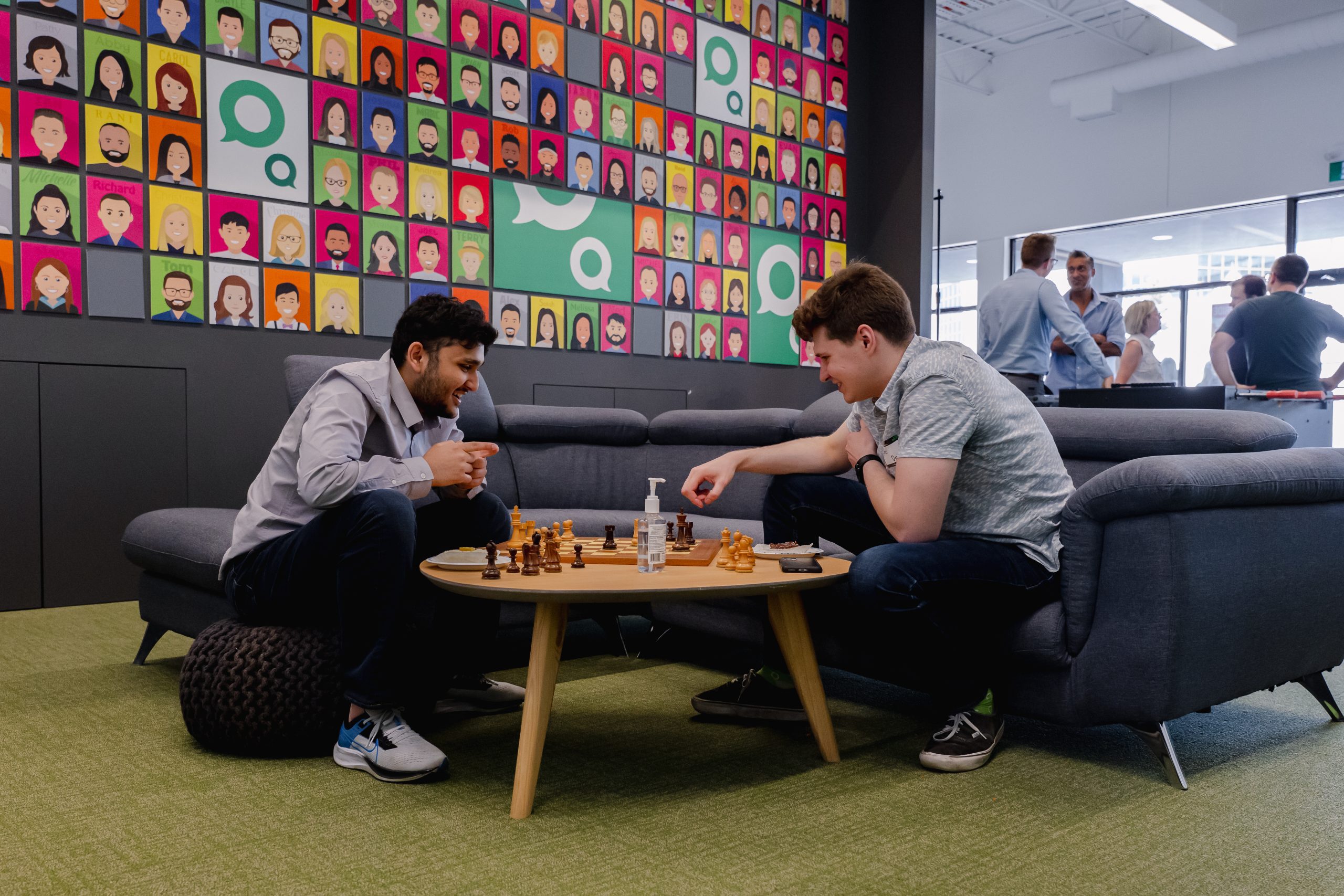 Two men playing chess on a coffee table in a modern office lounge area. A wall in the background features a grid of colorful illustrated employee portraits and Axonify speech bubble logos.