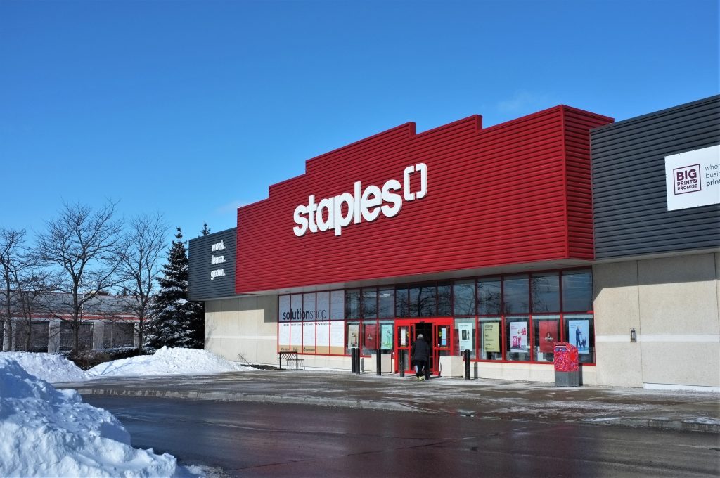 The exterior storefront of a Staples retail building featuring its signature red corrugated facade and white logo. Large display windows line the front entrance, including a sign for "solutionshop." Large piles of plowed snow sit along the wet asphalt parking lot in the foreground under a clear blue sky.